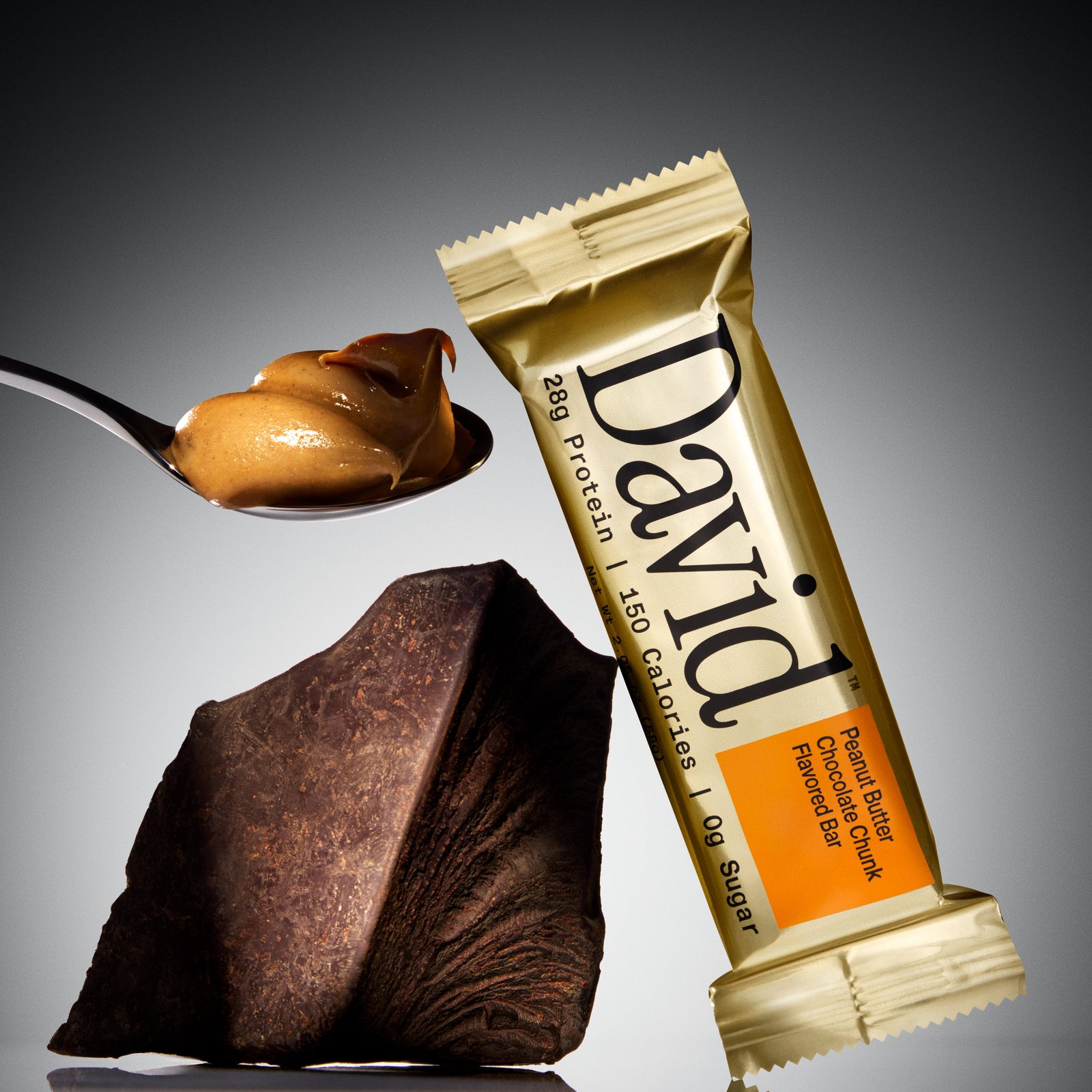 David protein bar with a spoonful of peanut butter and a piece of chocolate on a gray background