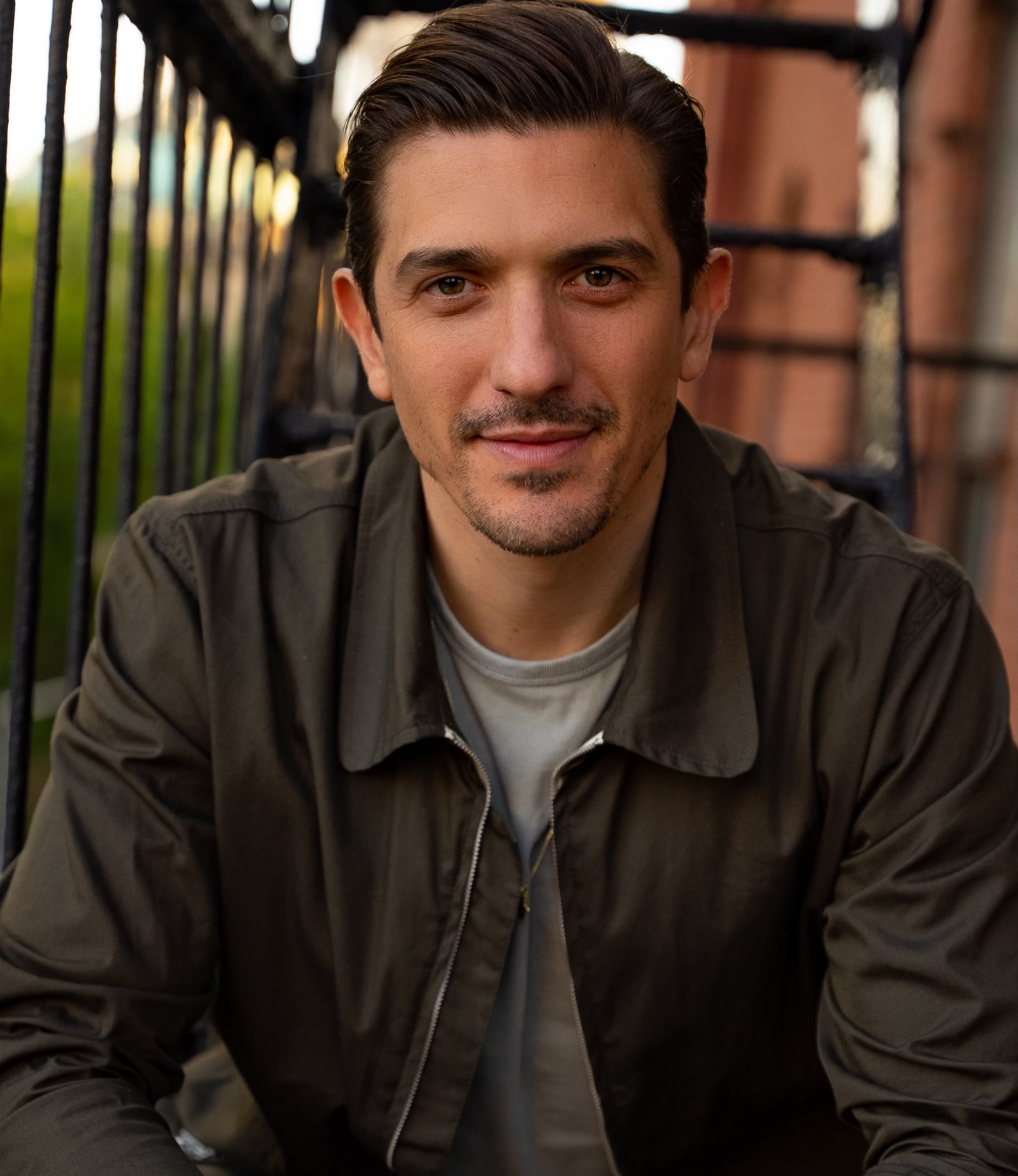Portrait of Andrew Schulz, a man in a grey shirt and a dark grey jacket on outdoor stairs