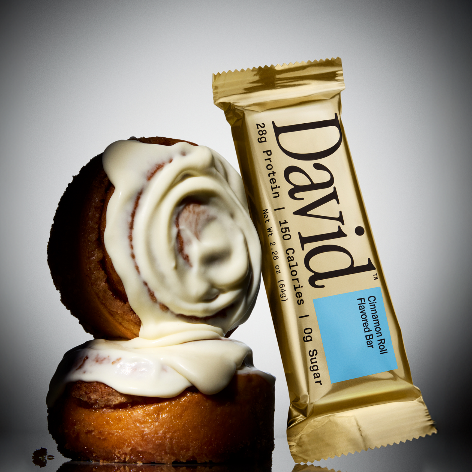 Cinnamon roll with cream cheese frosting and a David Cinnamon Roll flavored protein bar on a gray background