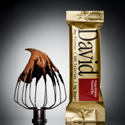 Fudge brownie flavored David protein bar next to a whisk with chocolate frosting on a gray background