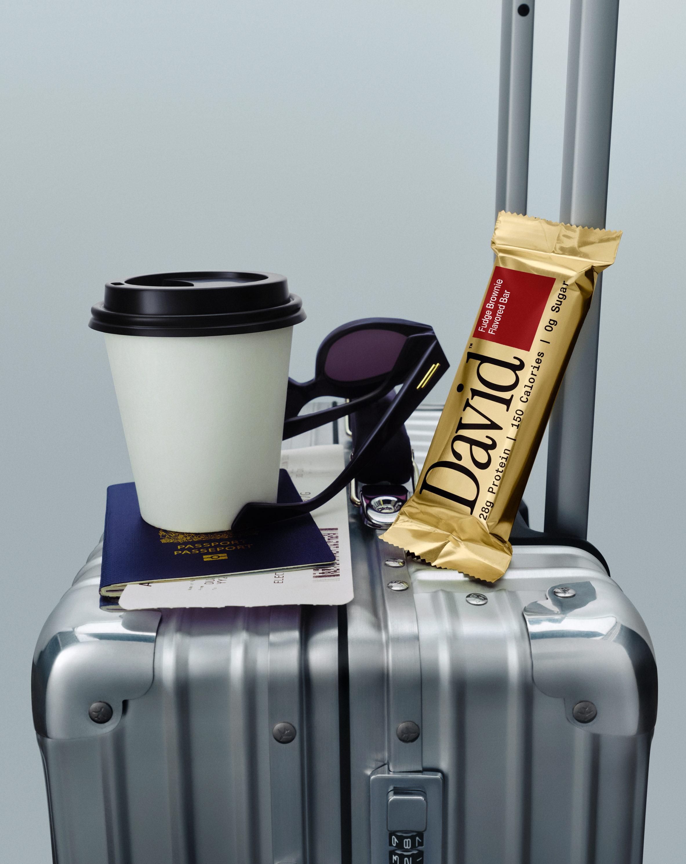 David Gold Fudge Brownie protein bar and coffee on a suitcase with travel accessories.