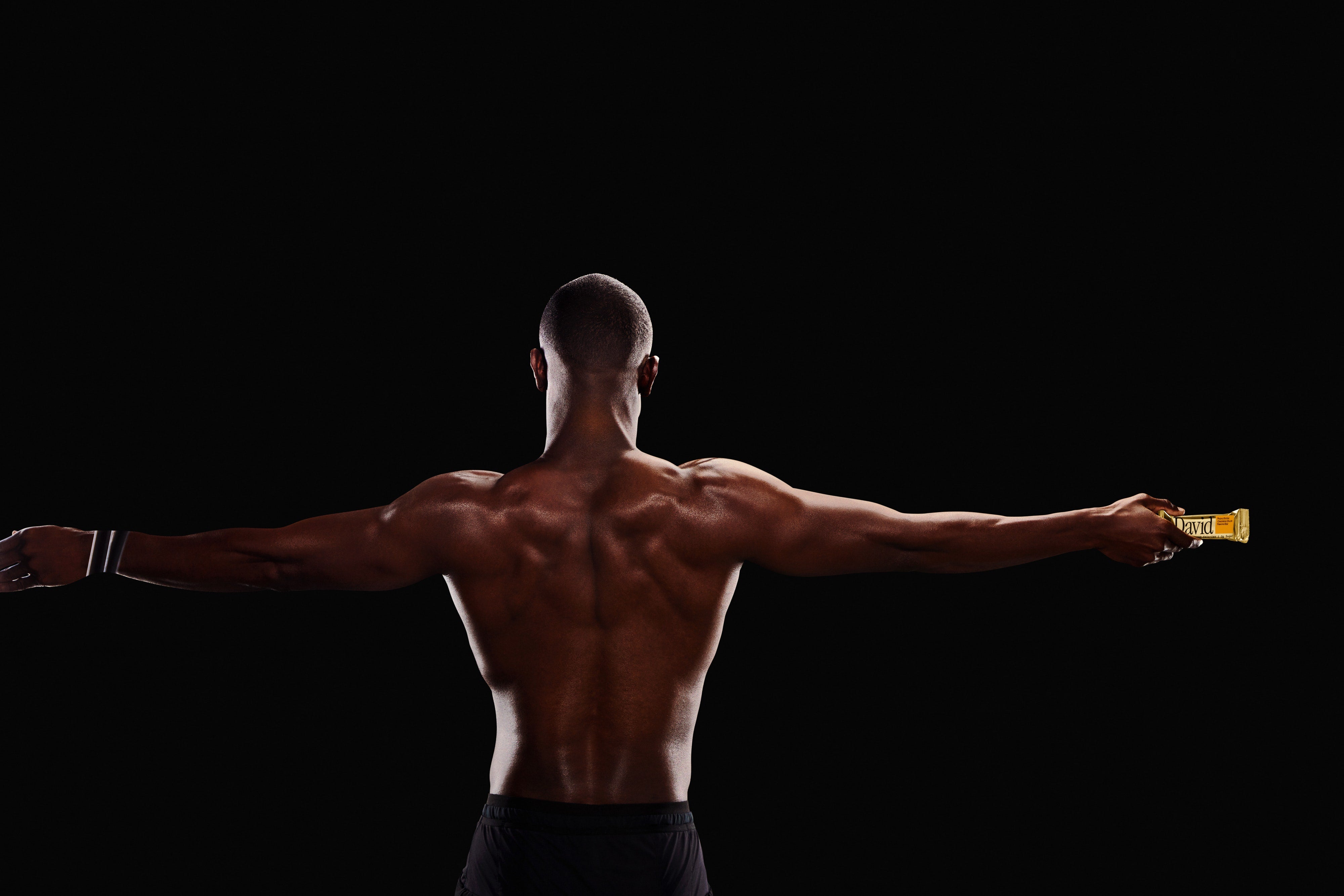 Man with a muscular back holding a gold protein bar against a black background