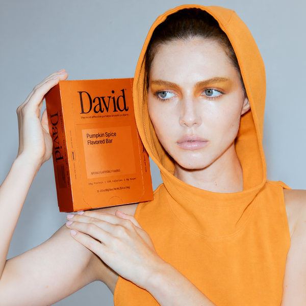 Woman in an orange hooded shirt holding David Pumpkin Spice protein bar box on her shoulder