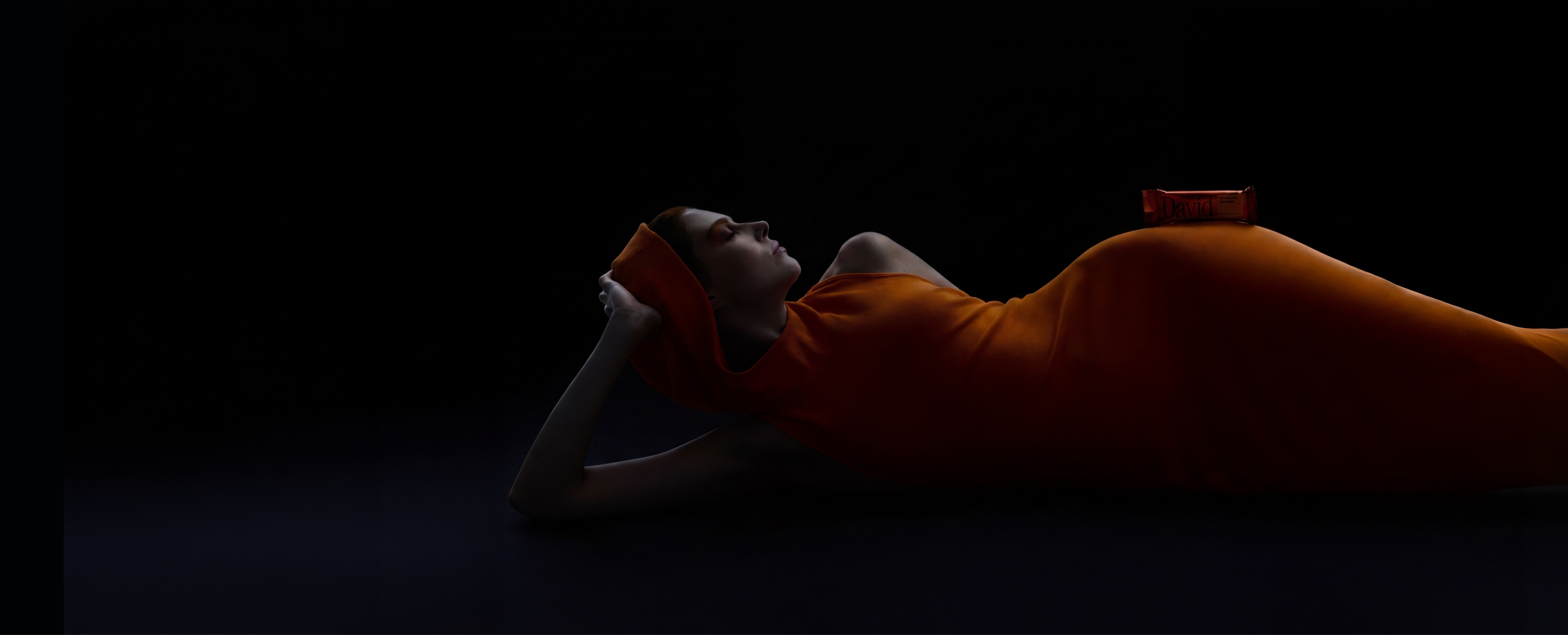 Model laying on her side in a hooded orange dress with a David Pumpkin Spice protein bar on her hip